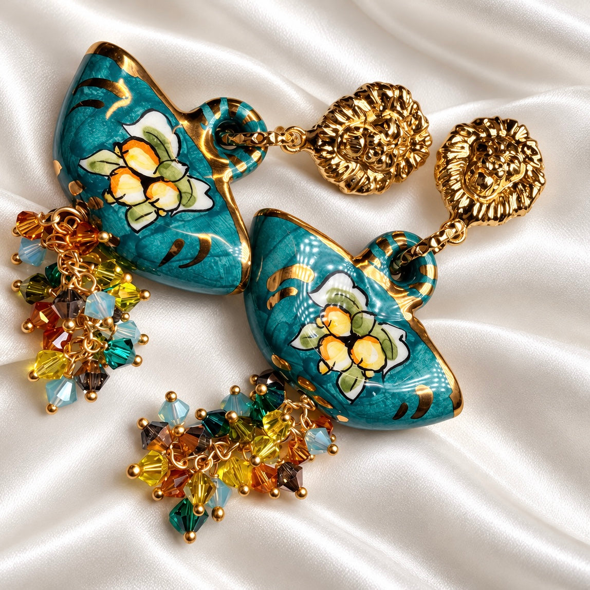 Angled view of turquoise Caltagirone ceramic coffa earrings with gold-tone lion studs and multicolored Swarovski crystals on white satin.