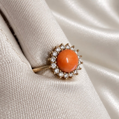 Close-up of a gold-tone ring with an orange cabochon surrounded by clear stones in a prong setting, on a white satin glove.