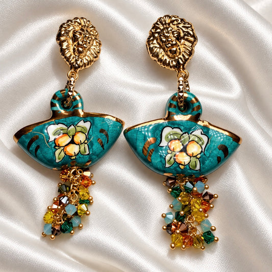Turquoise Caltagirone ceramic coffa earrings with floral motifs, gold-tone lion studs, and Swarovski crystal clusters on white satin.