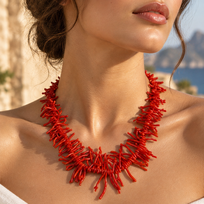 Close-up of a woman wearing a vivid red necklace with clustered coral-shaped branches against a sunlit coastal backdrop.