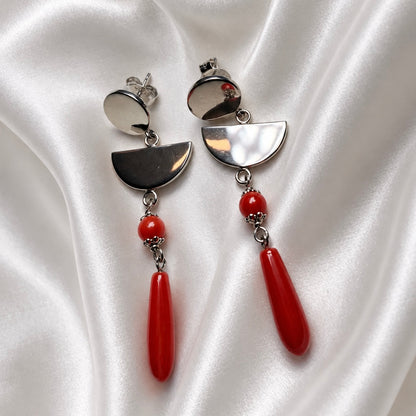 Angled view of Sardinian red coral and rhodium-plated silver earrings with polished semicircles and elongated red drops on white satin.
