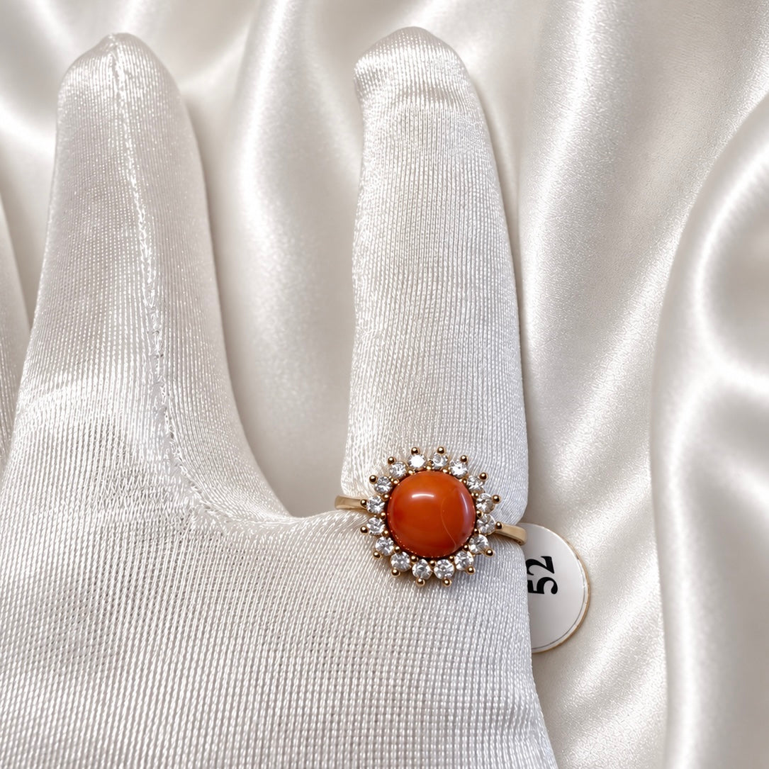 Gold-tone ring with a round orange-red cabochon and a halo of clear stones, displayed on a white satin glove.