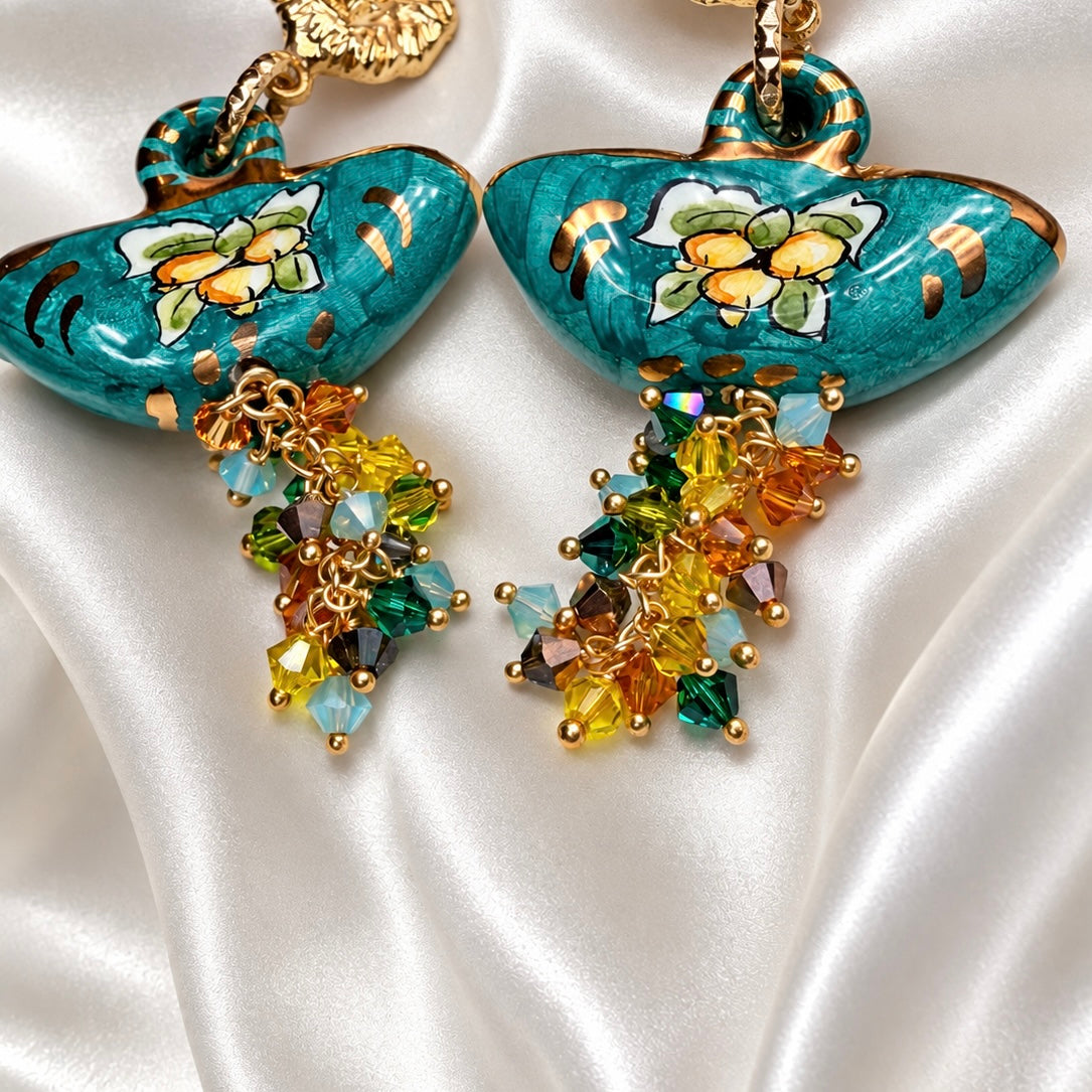 Close-up of turquoise Caltagirone ceramic coffa earrings with painted floral motifs, gold accents, and multicolored Swarovski crystal clusters.