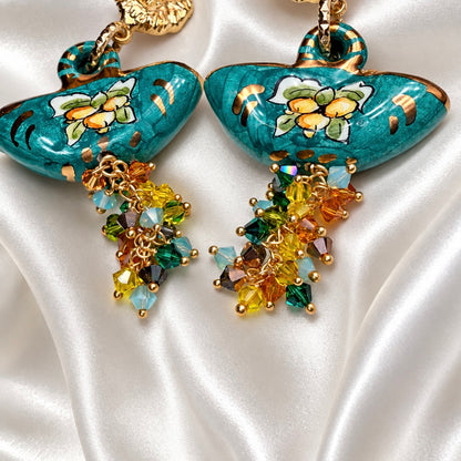 Close-up of turquoise Caltagirone ceramic coffa earrings with painted floral motifs, gold accents, and multicolored Swarovski crystal clusters.
