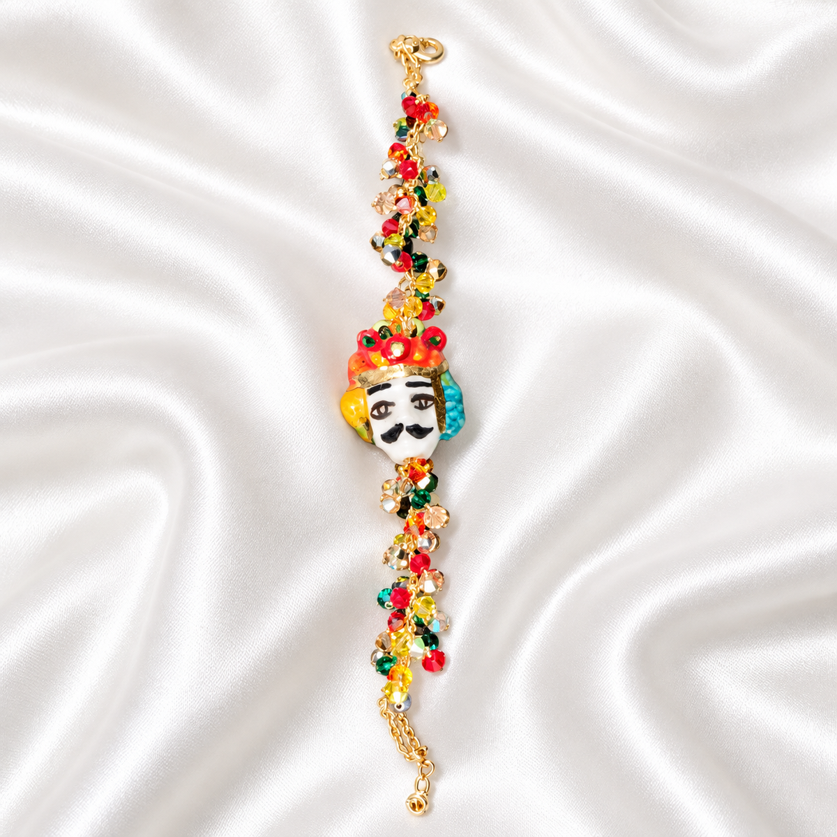 Full-length view of handmade Sicilian bracelet featuring a hand-painted ceramic Moor’s Head, multicolored Swarovski crystals and a gold-plated brass chain on white satin.

